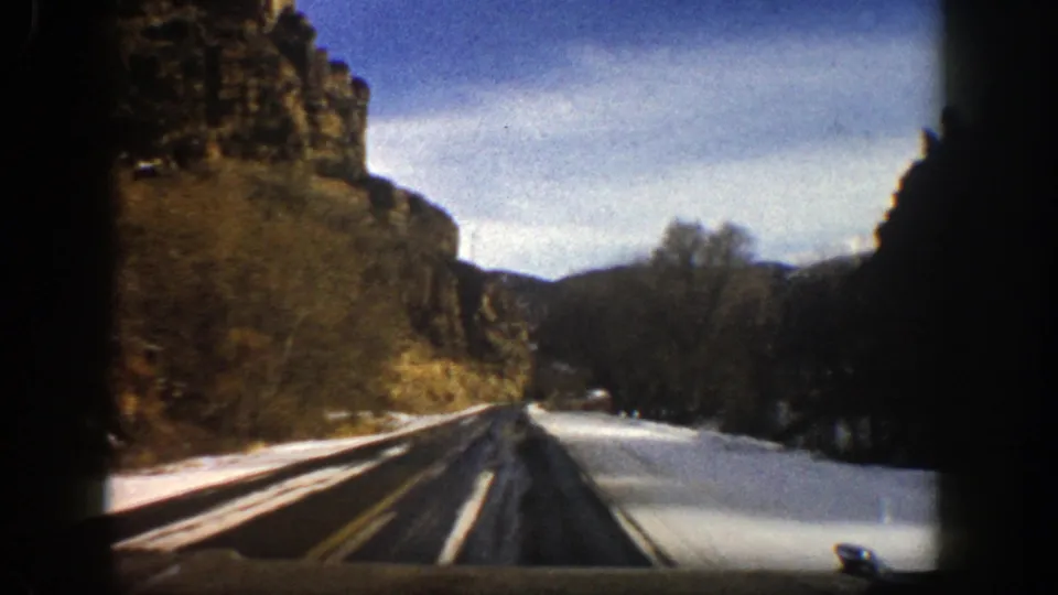 1961 Aspen-area road-trip frame showing a divided asphalt highway bordered by snow, cliffs, bare trees, and mountain slopes.