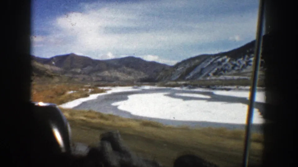 1961 Aspen-area 8mm frame from a moving vehicle overlooking a frozen or snow-covered water body with mountains beyond.