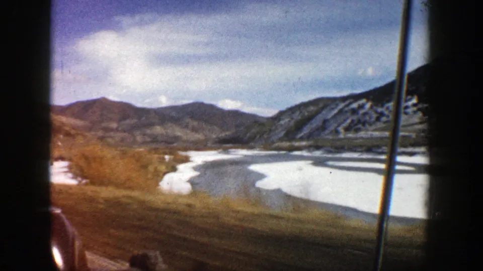 1961 Aspen-area travel frame looking past a car window toward a snowy lake or river bend, bare trees, and mountain ridges.