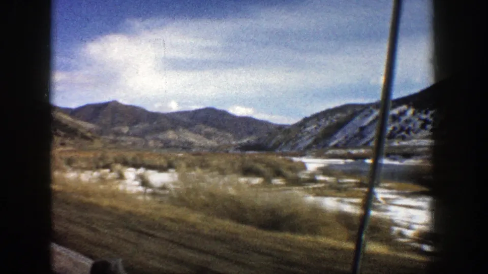 1961 Colorado road view with dry grass, scattered snow, a waterway, and distant hills seen through car-window framing.