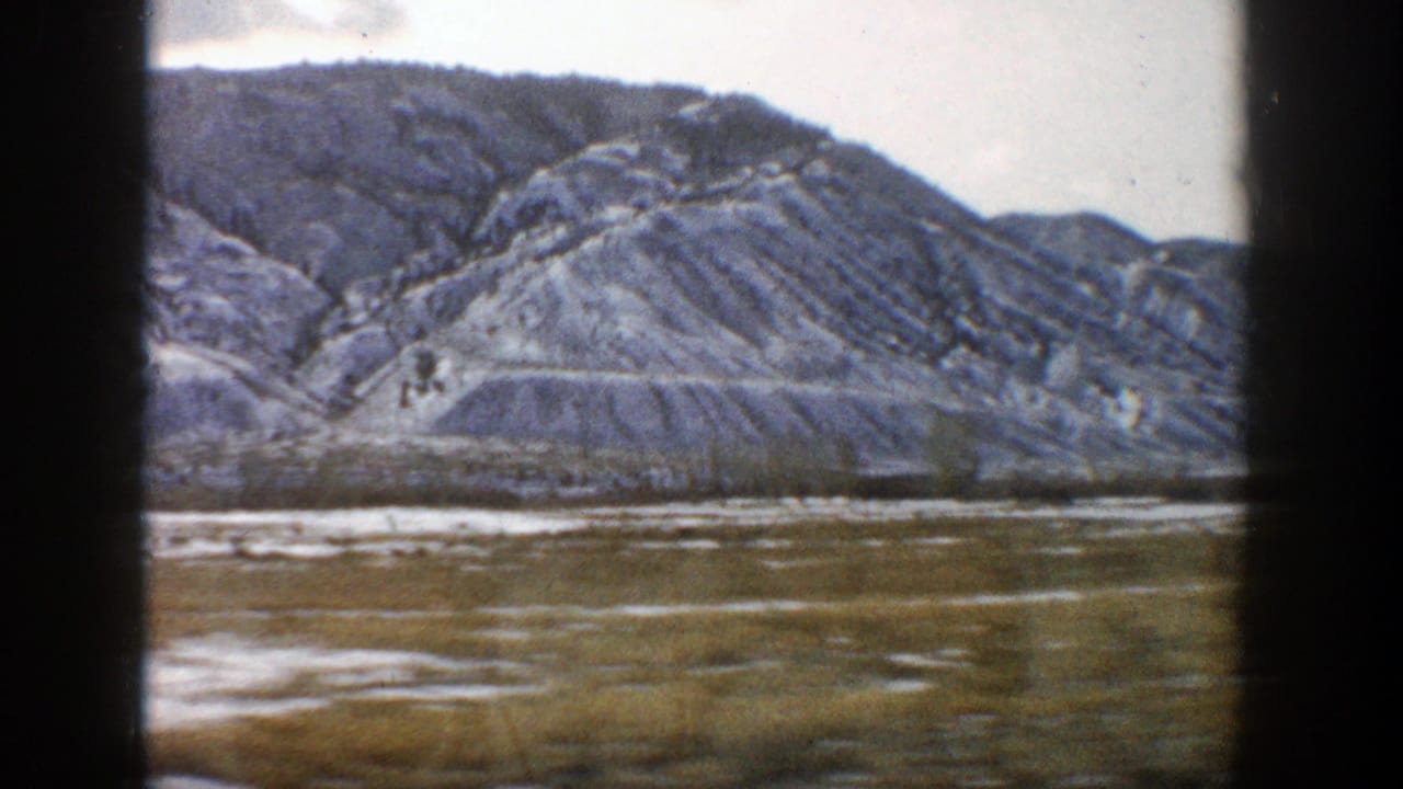 Snow-streaked rocky mountains seen past a dark train-window frame.