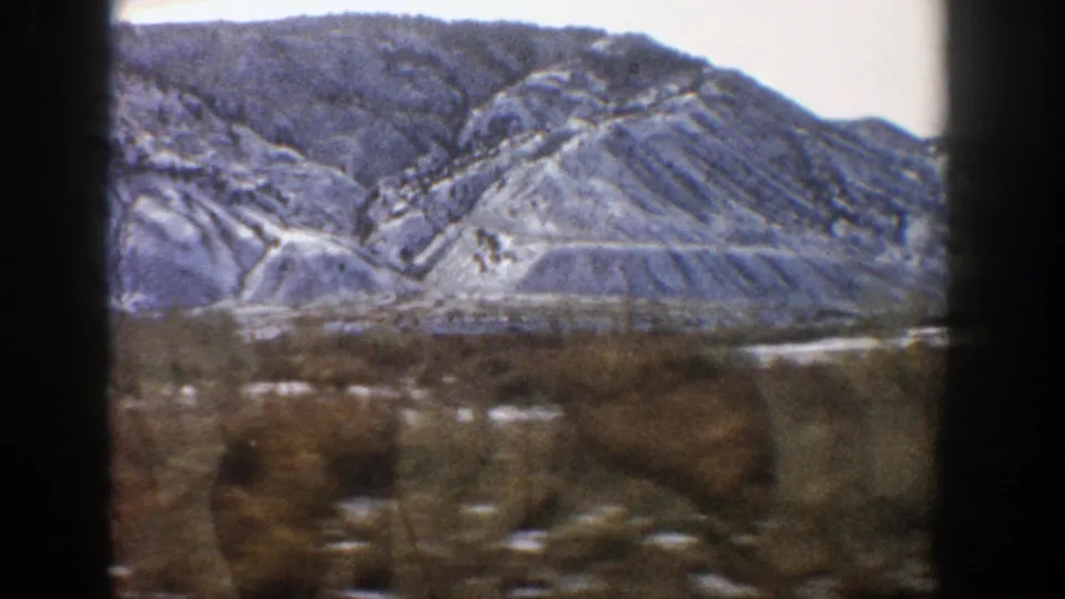 Final 1961 Aspen, Colorado frame showing a snow-dusted mountain slope beyond brown brush, with 8mm grain and motion blur.