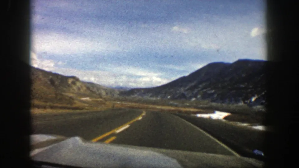 1961 home-movie frame of an empty Aspen mountain road, bordered by snow patches and rocky hills.