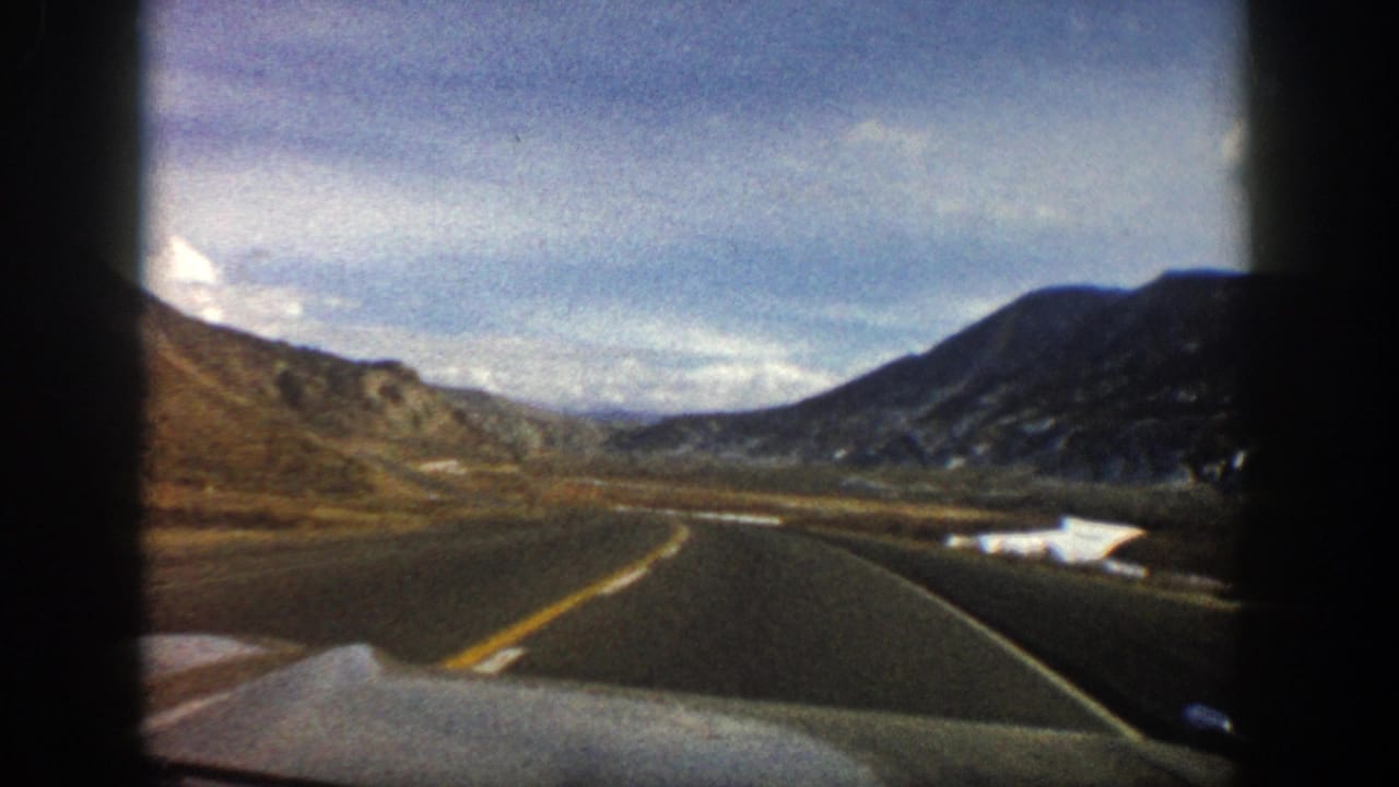 View through a car windshield of a curving mountain road with hills and cloudy sky.