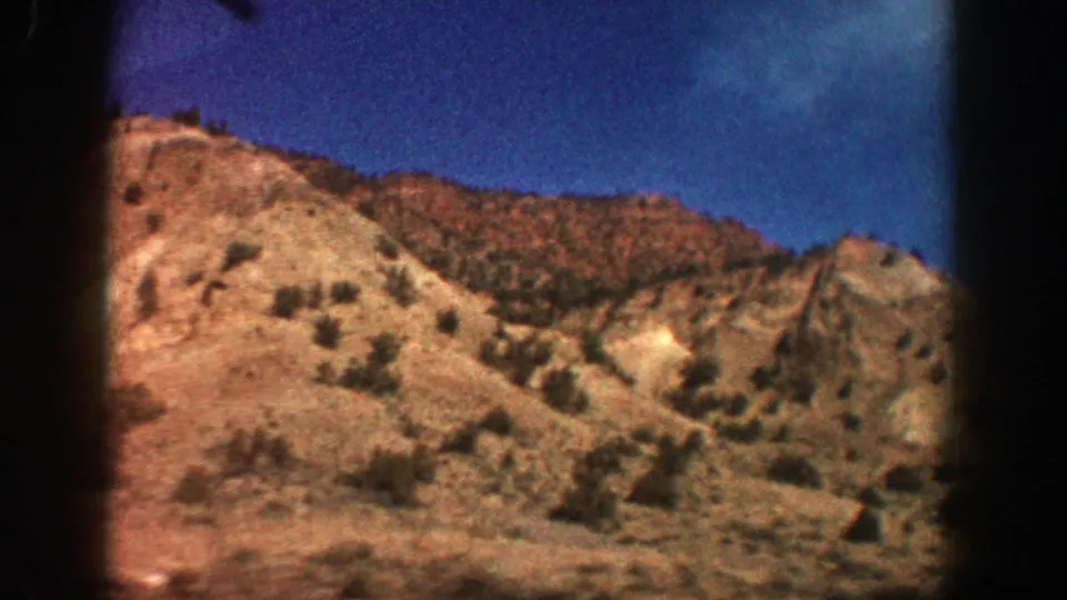 1961 8mm frame from Aspen showing a closer arid hillside view with brush, rocky ridges, and open blue sky.