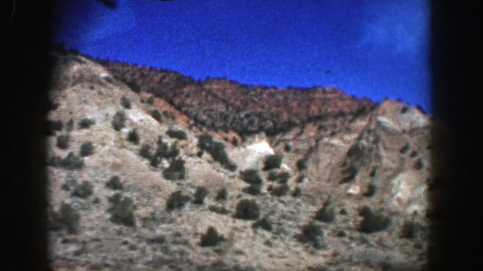 Aspen, Colorado home-movie frame showing rugged tan-gray hillside terrain, sparse shrubs, and bright blue sky.