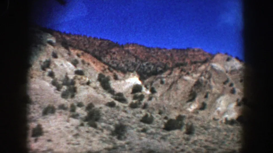 1961 Aspen, Colorado road-trip frame showing a dry rocky hillside, scrub vegetation, and blue sky through dark film edges.