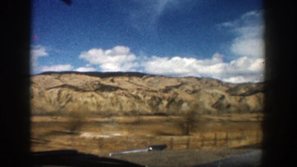 1961 road-trip view from a vehicle showing open dry terrain, low hills, scattered clouds, and a dark car edge at the bottom.