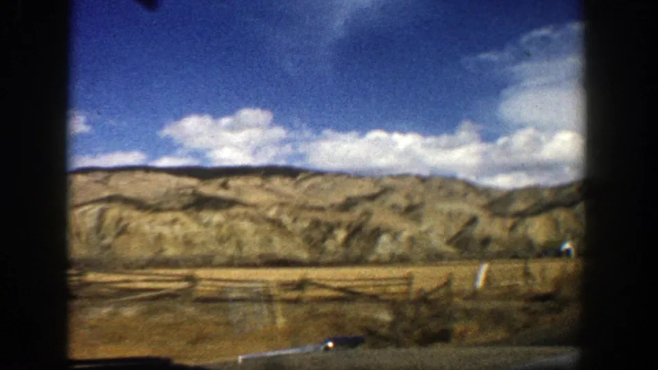 Aspen, Colorado home-movie frame with dry roadside ground, low ridgelines, scattered clouds, and the edge of a vehicle visible below.