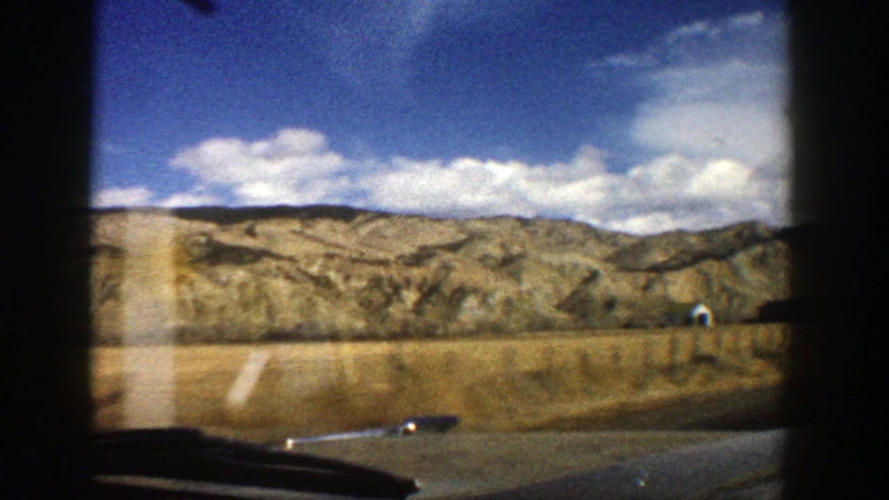 1961 8mm home-movie view near Aspen, Colorado, showing dry fields, rugged hills, clouds, and a road edge through a car window.