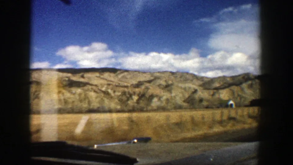 1961 8mm home-movie view near Aspen, Colorado, showing dry fields, rugged hills, clouds, and a road edge through a car window.