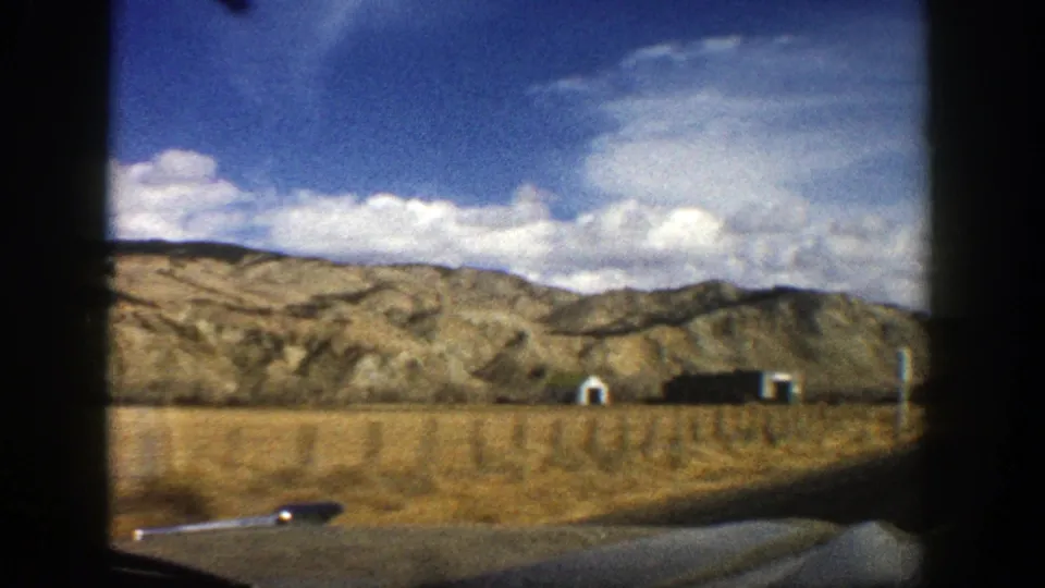 1961 Aspen, Colorado road view from inside a moving car, with dry fields, low buildings, fence posts, and rugged hills under clouds.