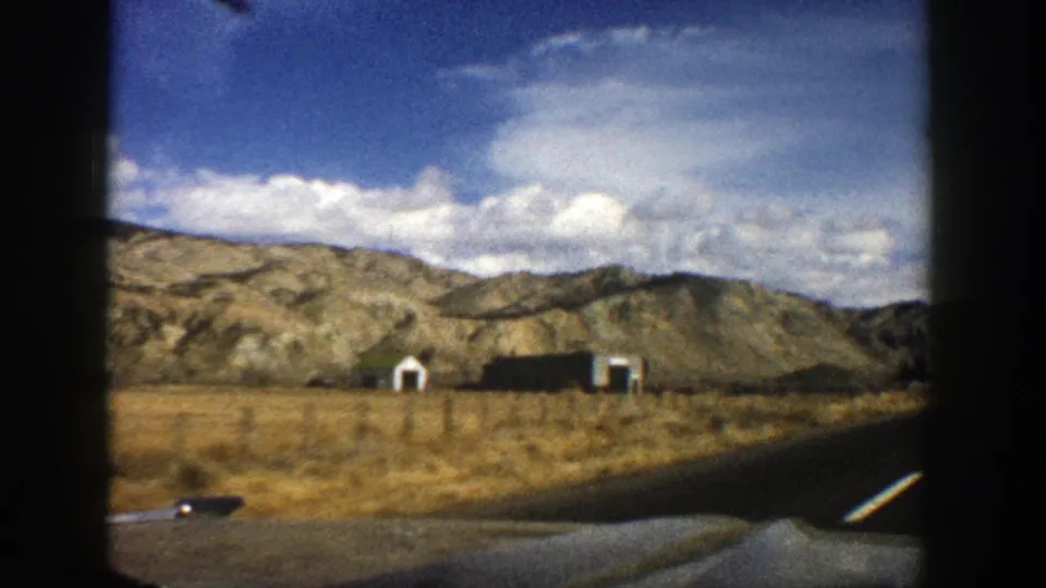 1961 8mm roadside view near Aspen, Colorado, showing small buildings, a fence line, dry grass, hills, and a blue sky.