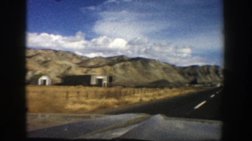 1961 Aspen, Colorado home-movie frame showing a paved road, car hood edge, dry roadside land, small buildings, and hills under clouds.