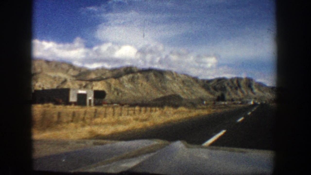 Paved road seen over a car hood with dry grass, roadside buildings, rocky hills, and clouds.