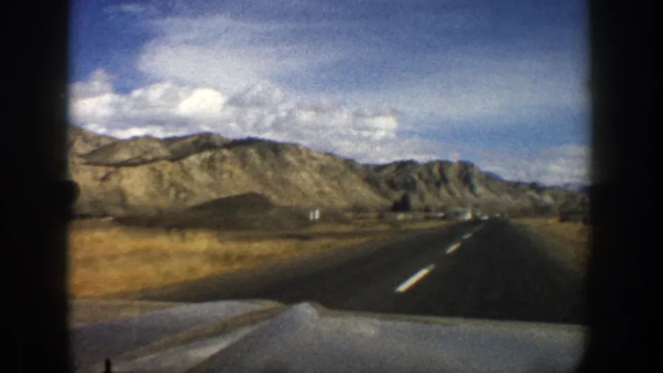 Forward view from a moving car on a rural Aspen, Colorado road in 1961, with dry valley ground and rocky hills ahead.