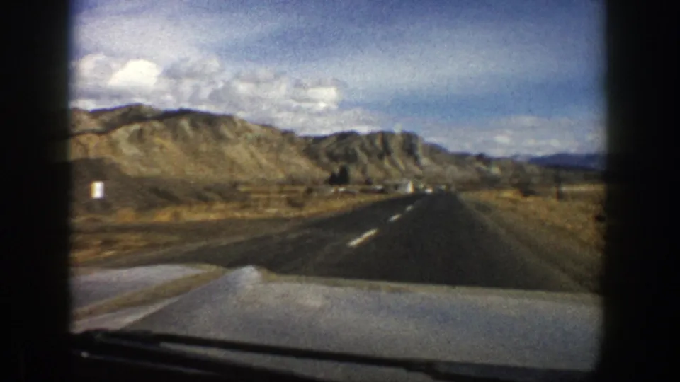 1961 Aspen-area road view from inside a car, showing a paved lane, dry roadside land, hills, and small distant roadside forms.