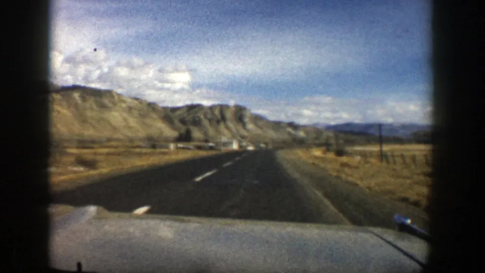 Final 1961 Aspen, Colorado frame shows the road bending through open dry ground, with fence posts, low buildings, and rocky hills beyond.