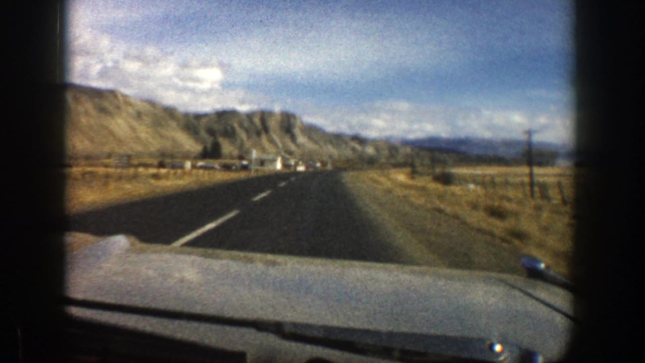 Forward view from a car on a two-lane road toward tan hills under a blue sky.