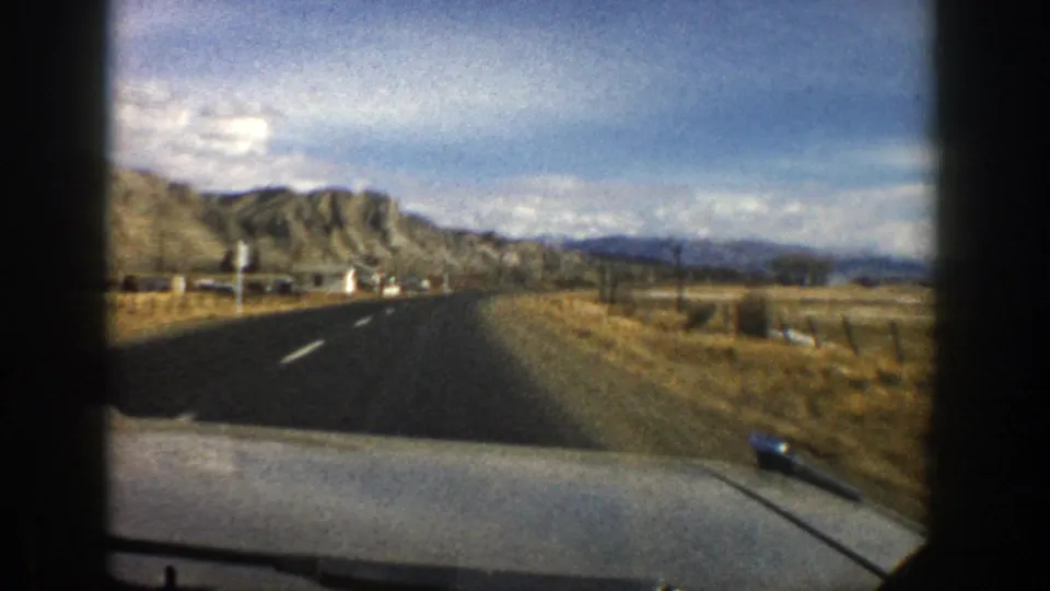 1961 Colorado road-trip frame showing a paved road beside dry grassland and tan hills from inside a moving vehicle.