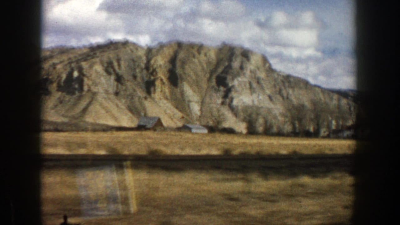 Mountain slope beyond dry open land with small buildings and dark film borders.