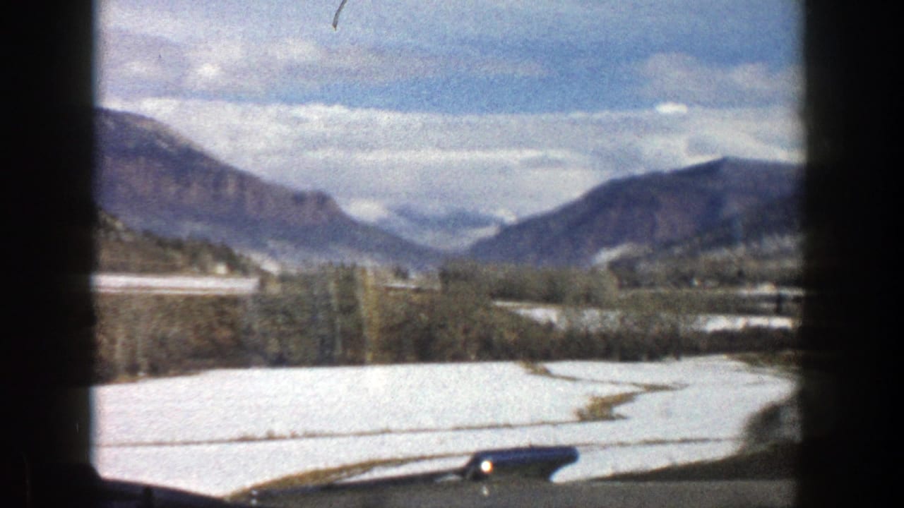 1961 Aspen, Colorado home-movie view of snow-dusted fields, leafless trees, mountains, and cloudy blue sky from a car window.