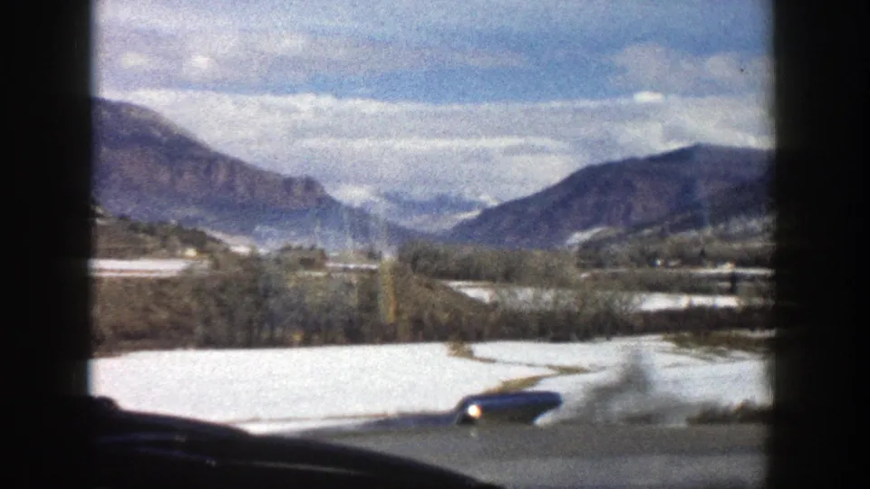Aspen, Colorado 8mm frame showing a mountain valley, pale snow patches, trees, and a cloudy sky from a moving vehicle in 1961.