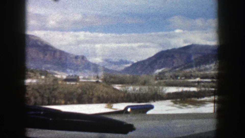 Home-movie frame from Aspen in 1961 with a passing roadside foreground, snowy field, bare trees, and distant mountain ridges.
