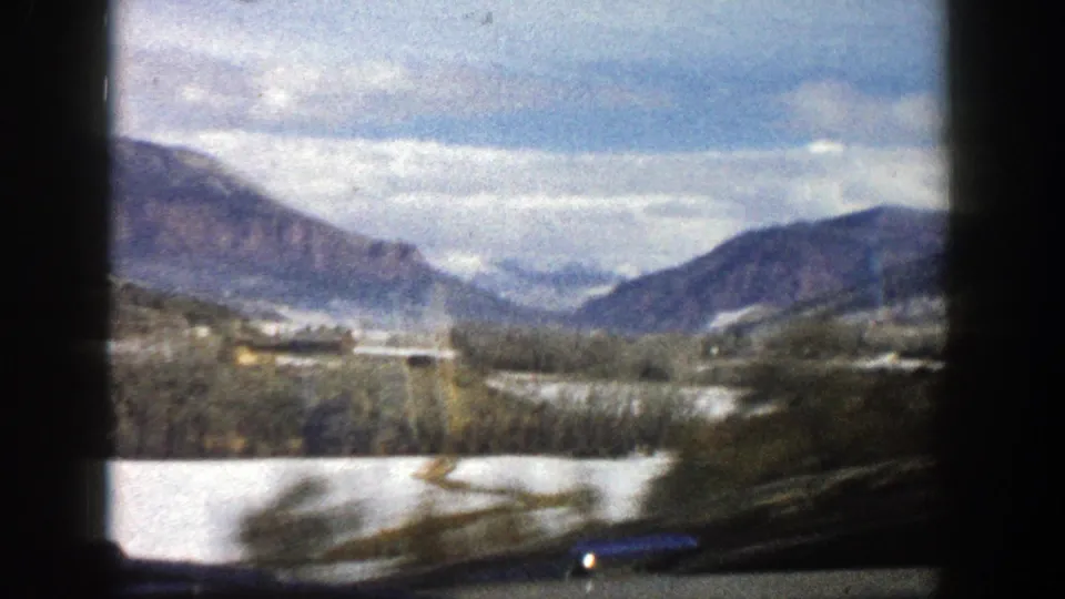 1961 Aspen, Colorado travel frame with motion-blurred roadside foreground, snow fields, tree line, and mountains under a cloudy sky.