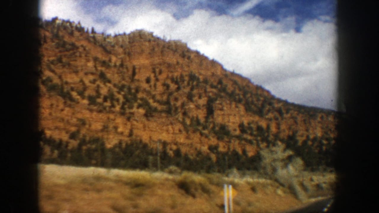 Curving mountain road beside a red-brown hillside and evergreen trees under clouds.