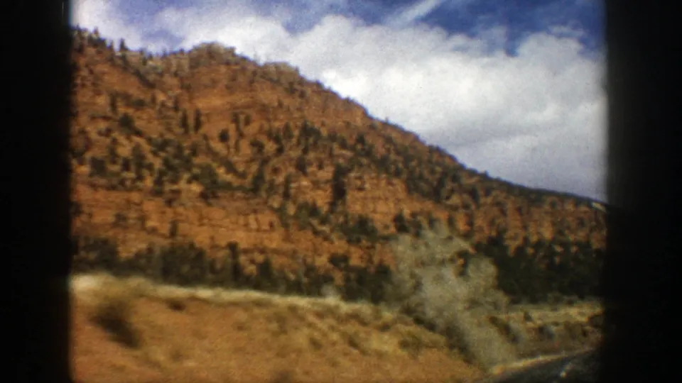 Passenger-view 8mm frame near Aspen, Colorado, with a red-brown hillside, scattered evergreens, and roadside vegetation.