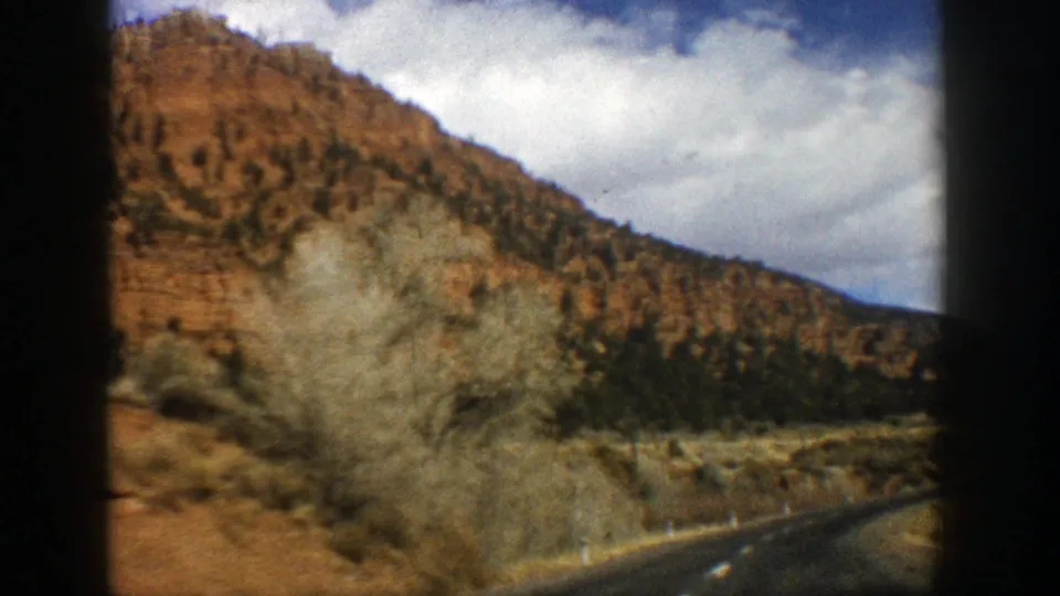 1961 Colorado road-travel frame showing dry brush, a paved road edge, and a red-brown mountain ridge under a cloudy sky.