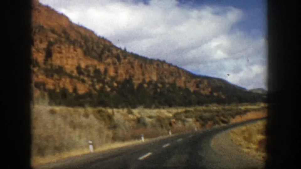 1961 Aspen, Colorado 8mm frame of a two-lane road bending through a mountain landscape with brush, evergreens, and clouds.