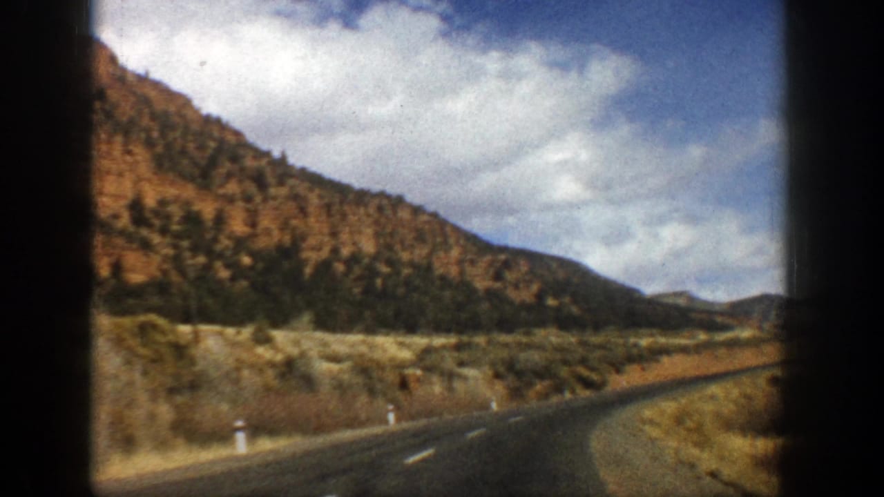 Curving mountain road bordered by dry scrub and hills under a partly cloudy sky.