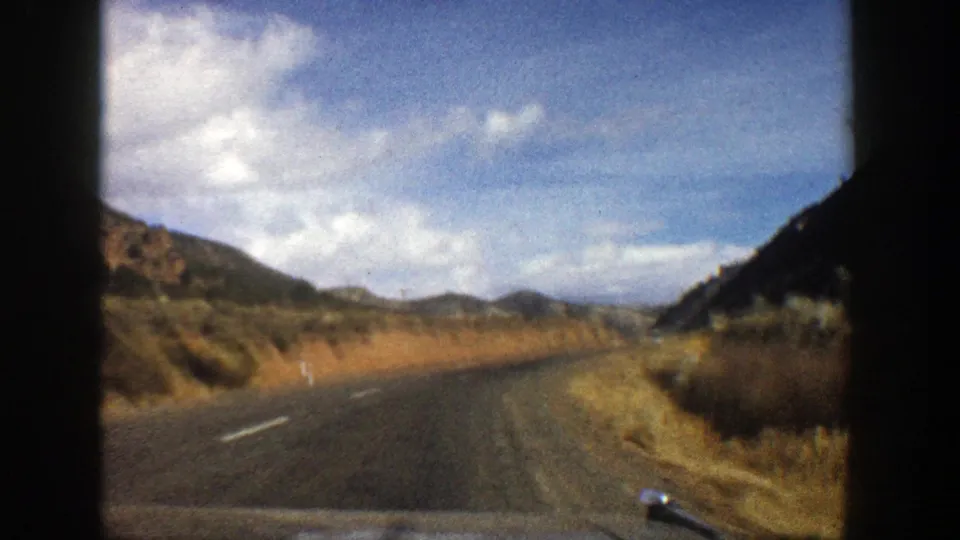 Car-mounted 8mm view of a quiet 1961 Aspen-area road approaching a bend between dry hills.