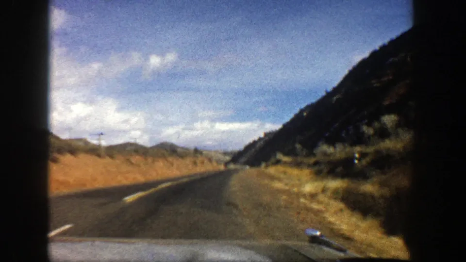 Late-frame 8mm view from a moving vehicle on a curving Aspen, Colorado road with hills and open sky visible.