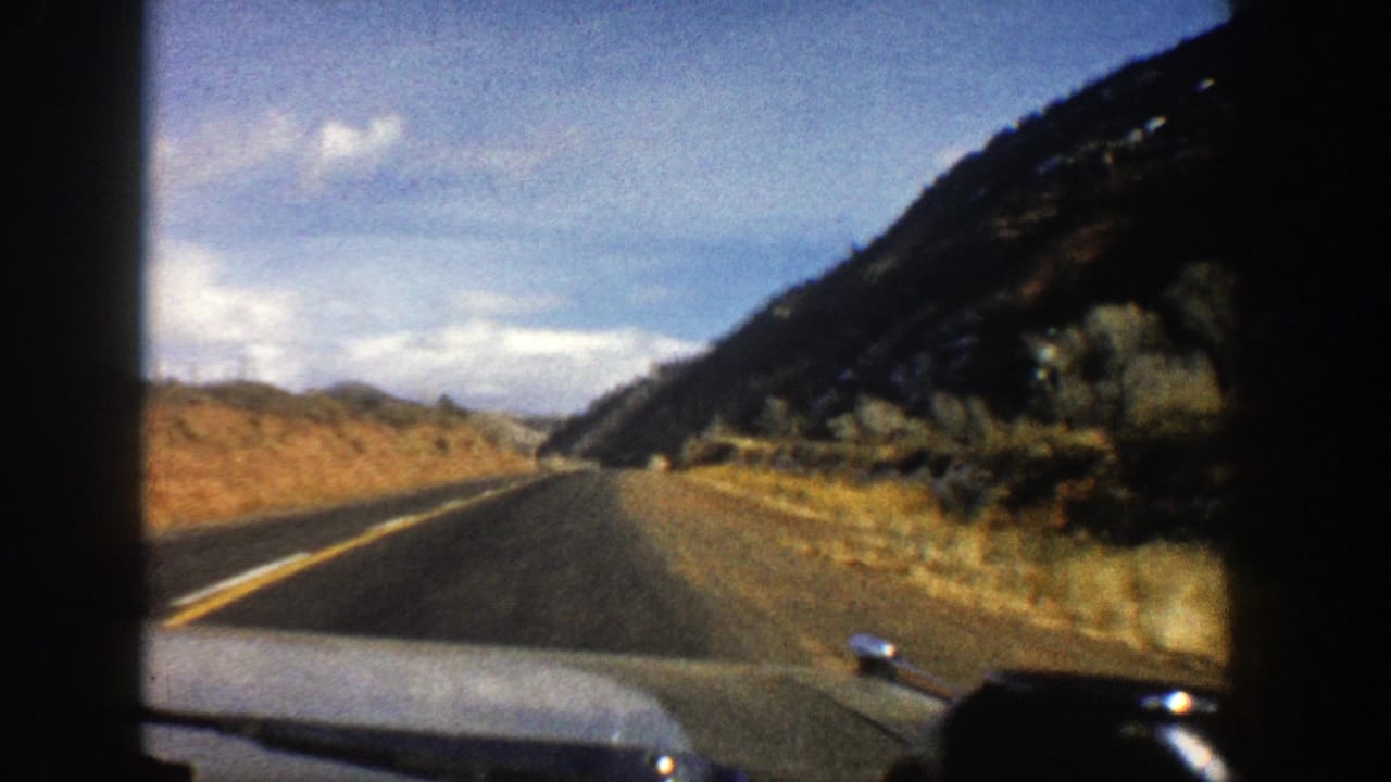 Two-lane mountain road seen over a car hood with rocky slopes and brush along the roadside.