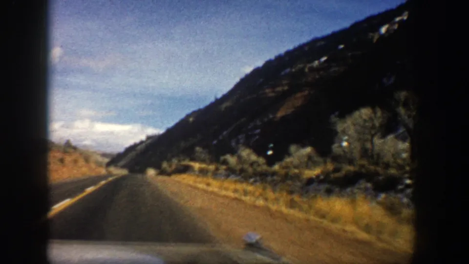 1961 Aspen-area driving view through the windshield, showing a paved road curving past mountain brush and a dark rocky slope.
