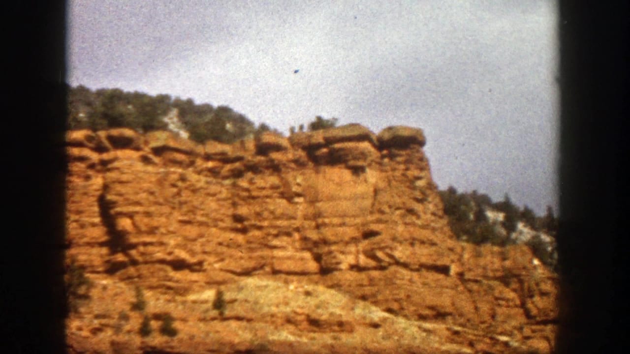 1961 Aspen, Colorado 8mm frame showing a reddish rock cliff, rounded capstones, green trees, and pale sky.