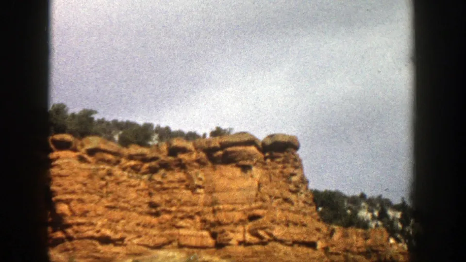 1961 Aspen, Colorado home-movie frame with a jagged reddish cliff, weathered ledges, hillside trees, and pale cloud cover.