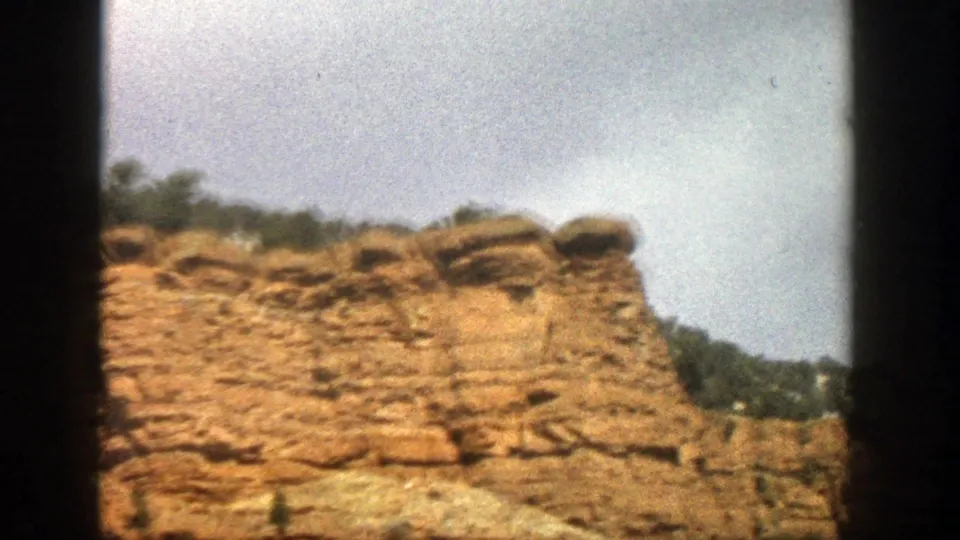 Soft 8mm view from 1961 Aspen, Colorado showing a layered orange cliff face, rounded rock top, and green trees near the ridge.