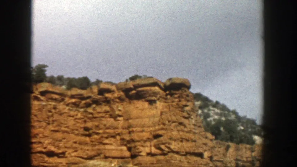 1961 Aspen, Colorado frame showing a broader view of the reddish cliff, capped ledges, forested hillside, and cloudy sky.