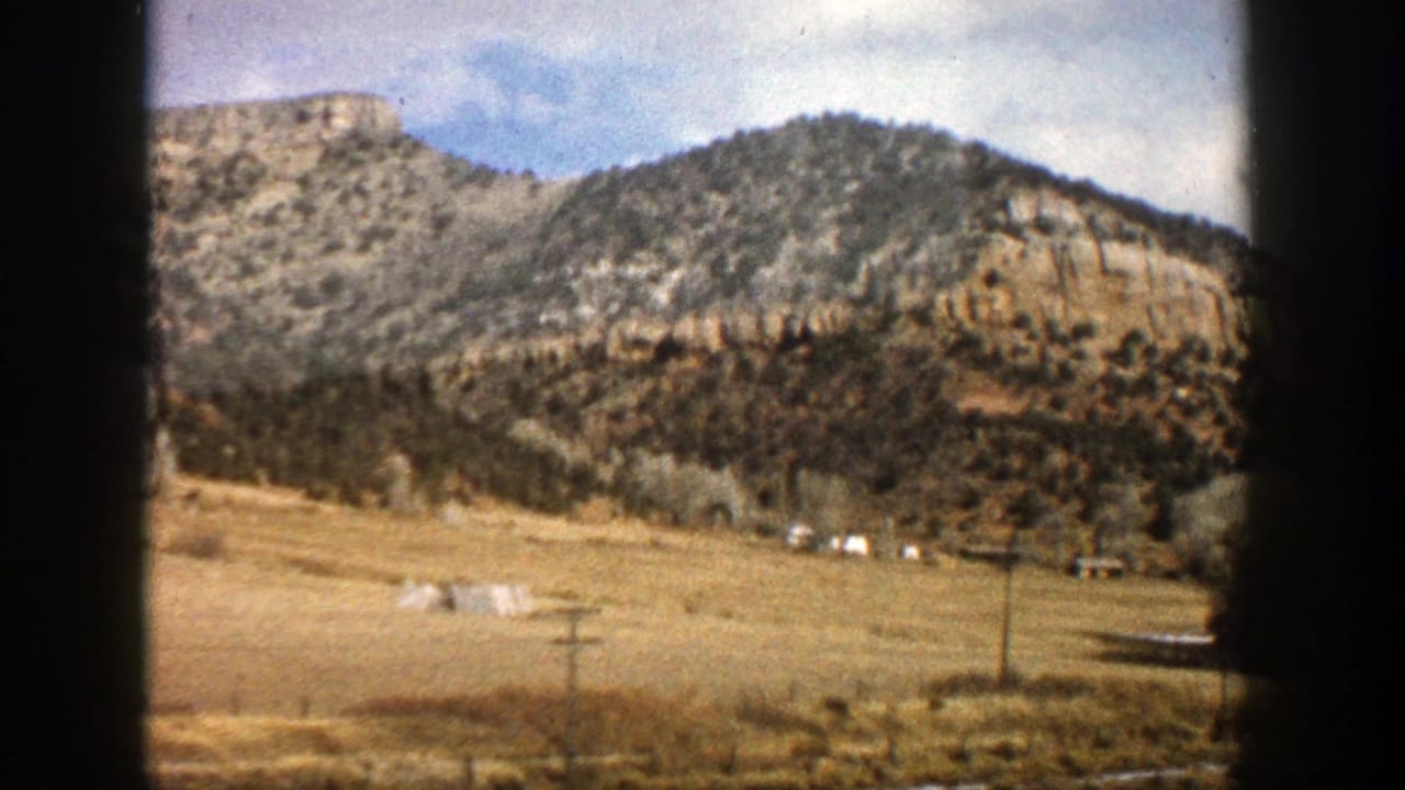 1961 Aspen, Colorado road-trip view through a vehicle window, showing sandy mountain slopes, dry valley ground, trees, and poles.
