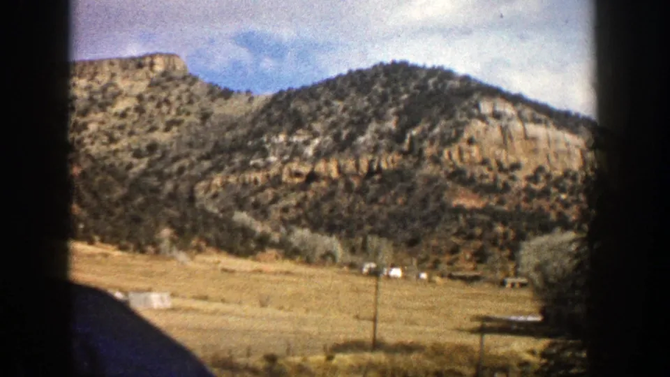 1961 Aspen, Colorado landscape seen from inside a vehicle, with a ridge, dry field, scattered trees, and distant structures.