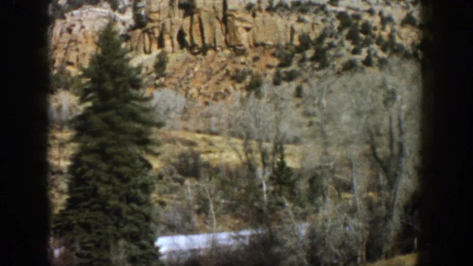 1961 Aspen-area roadside frame with layered rock, leafless aspen trees, an evergreen, and snow patches.