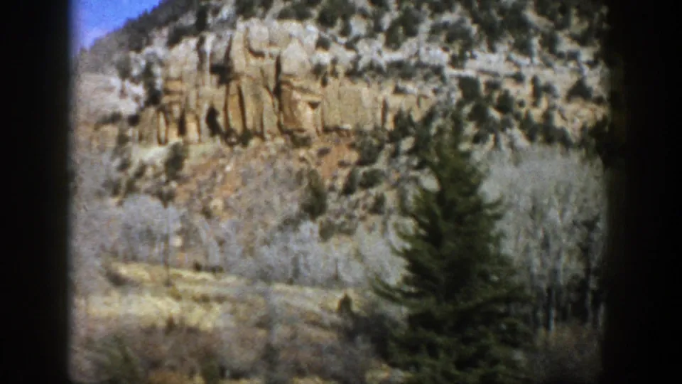 1961 Aspen, Colorado home-movie view of an evergreen and bare aspen trees against a tan cliff.