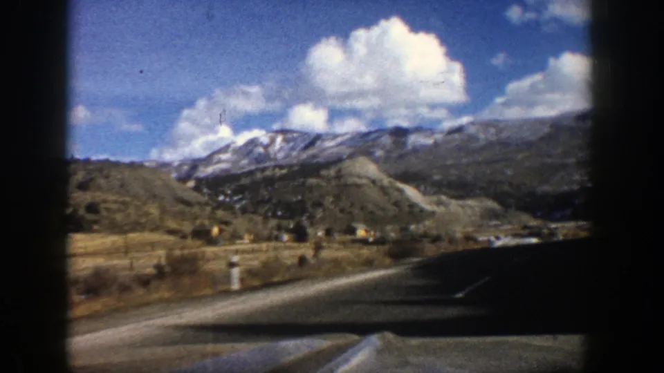1961 Aspen, Colorado 8mm view of a road, car window edge, rolling foothills, and distant snow-streaked mountains.