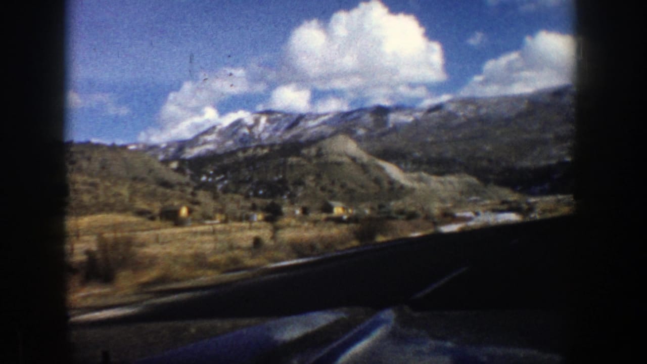 Mountain highway view with snow-dusted peaks, blue sky, clouds, and dark film borders.