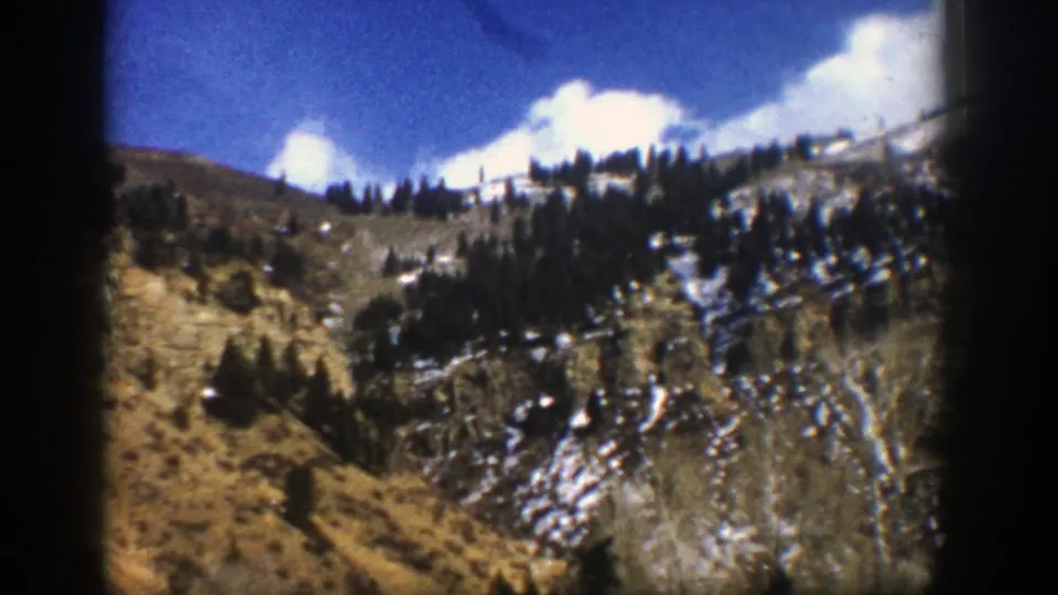 1961 Aspen, Colorado mountain slope seen from a moving vehicle, with evergreen trees, patchy snow, dry terrain, blue sky, and clouds.
