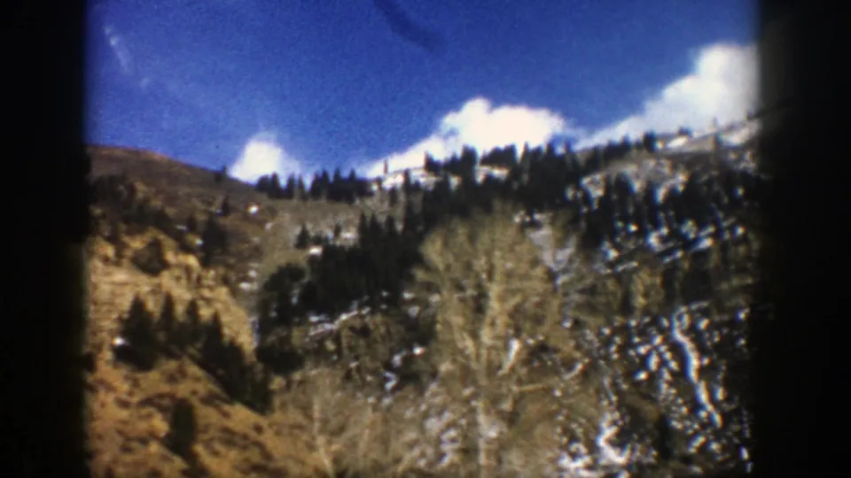 1961 Aspen, Colorado travel frame showing a rugged hillside, evergreen clusters, patchy snow, blue sky, clouds, and soft 8mm edges.